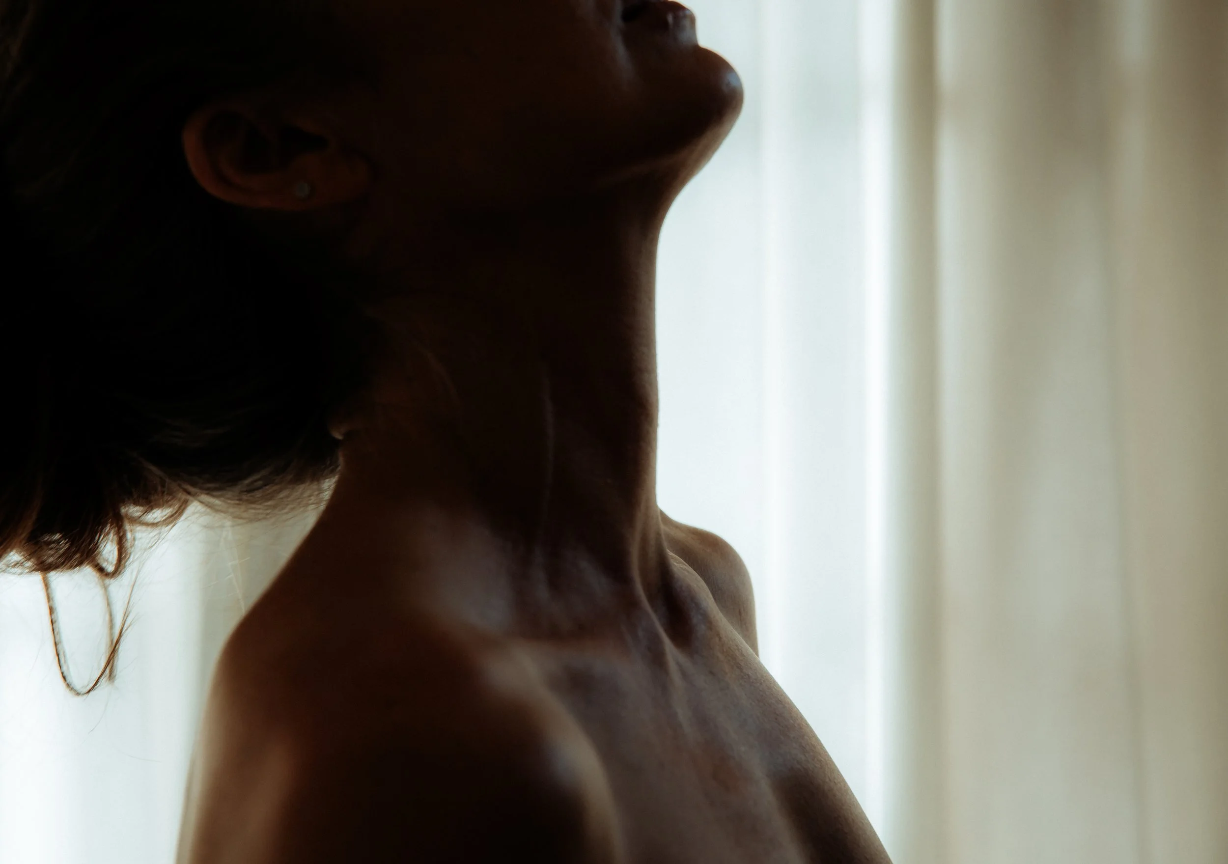 Close-up profile of a woman's face turned slightly upward with her eyes closed, illuminated by soft natural light, showing her neck, shoulder, and a small earring.