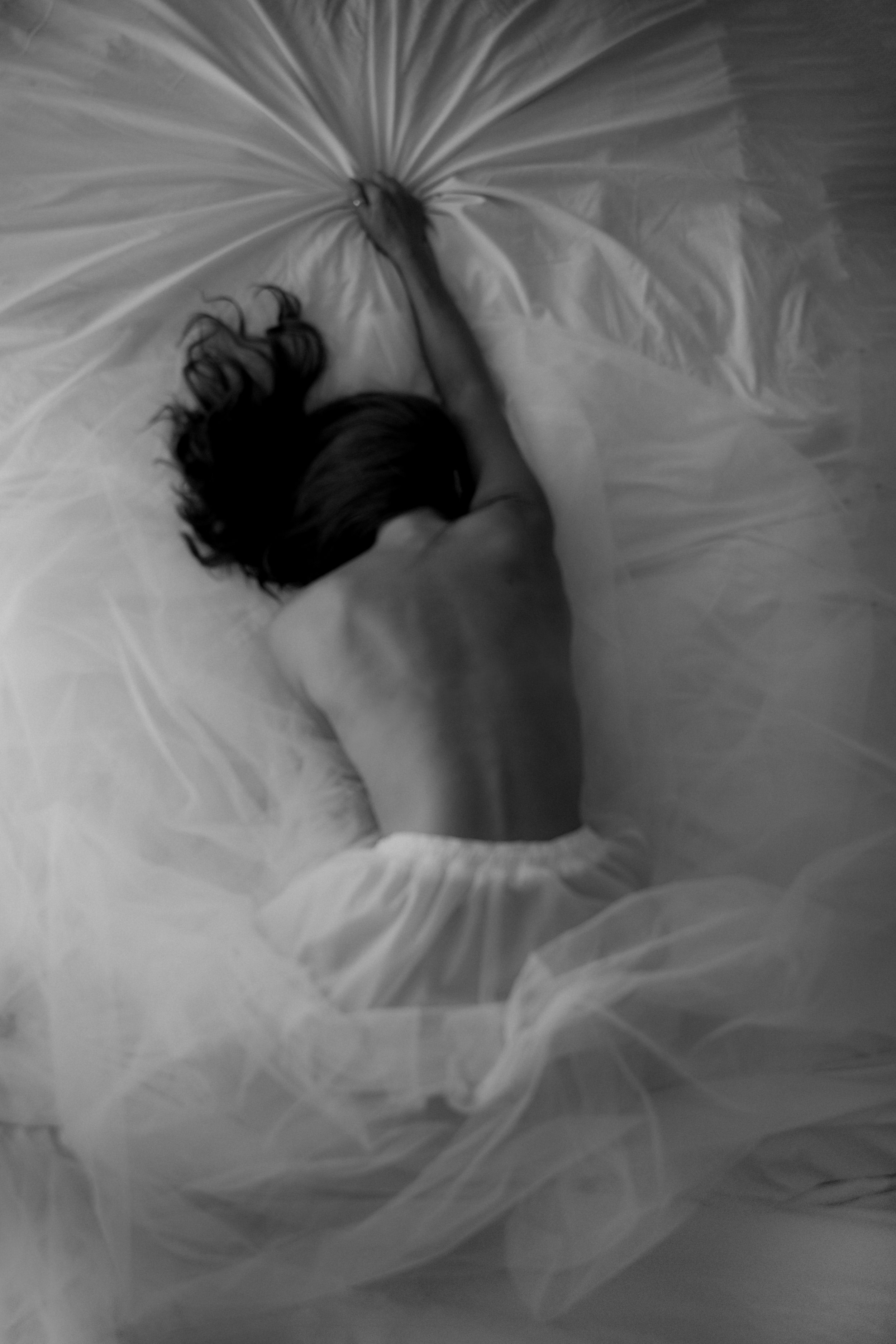 A person with long, curly hair is lying on the floor and touching a bed with their hand. The photo is in black and white.