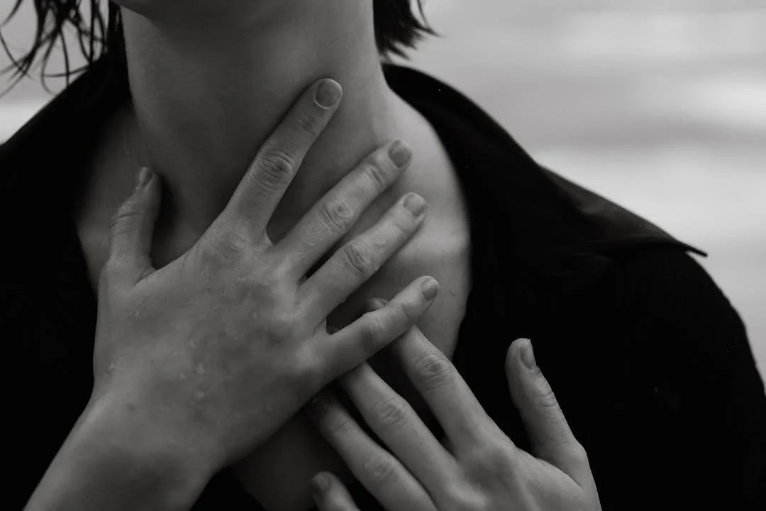 Black and white close-up of a person touching their throat with two hands, seemingly indicating discomfort or distress.