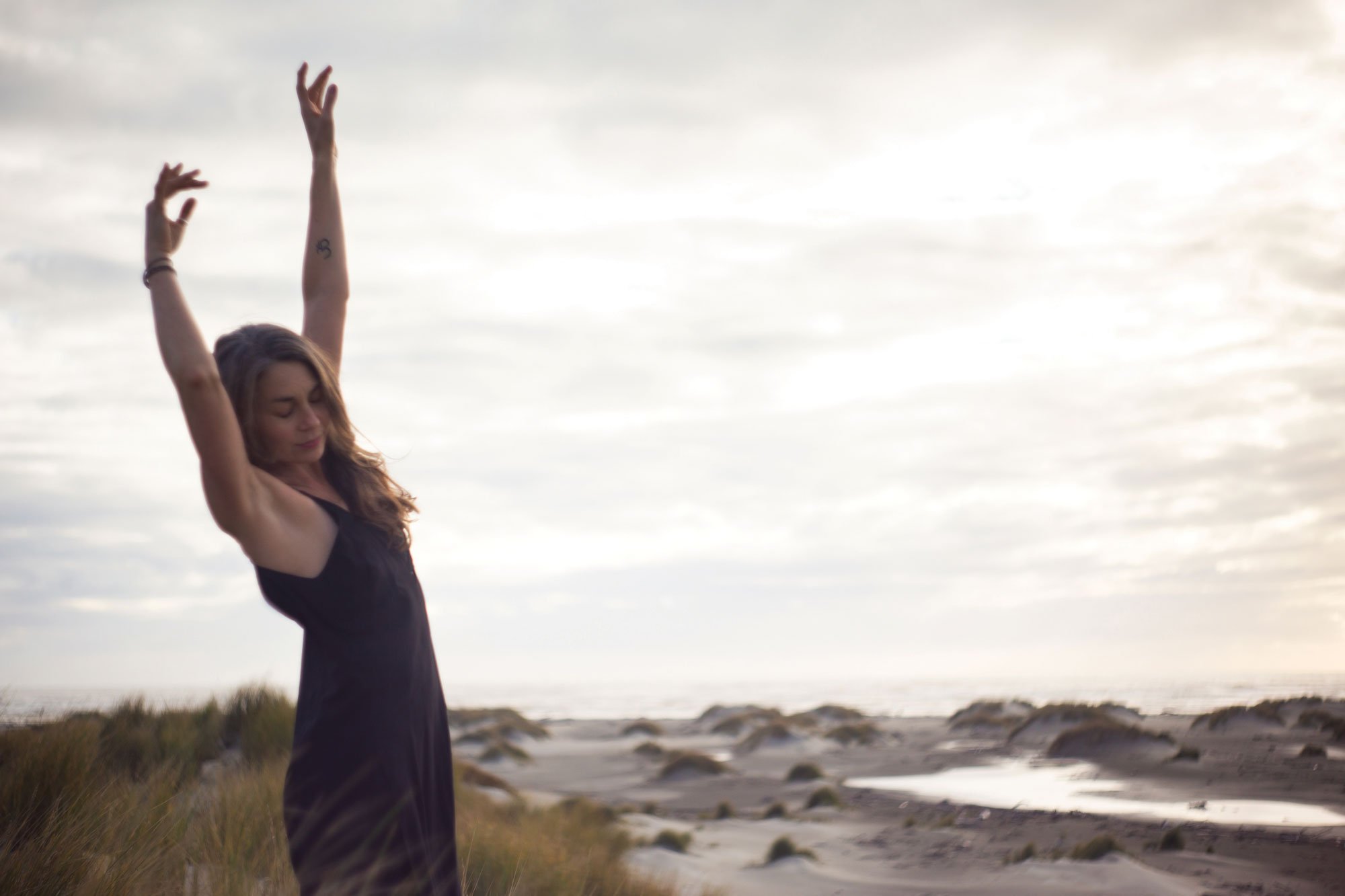 A woman with her arms raised in the air standing on a grassy area near a rocky shoreline, possibly watching the ocean or sunset.