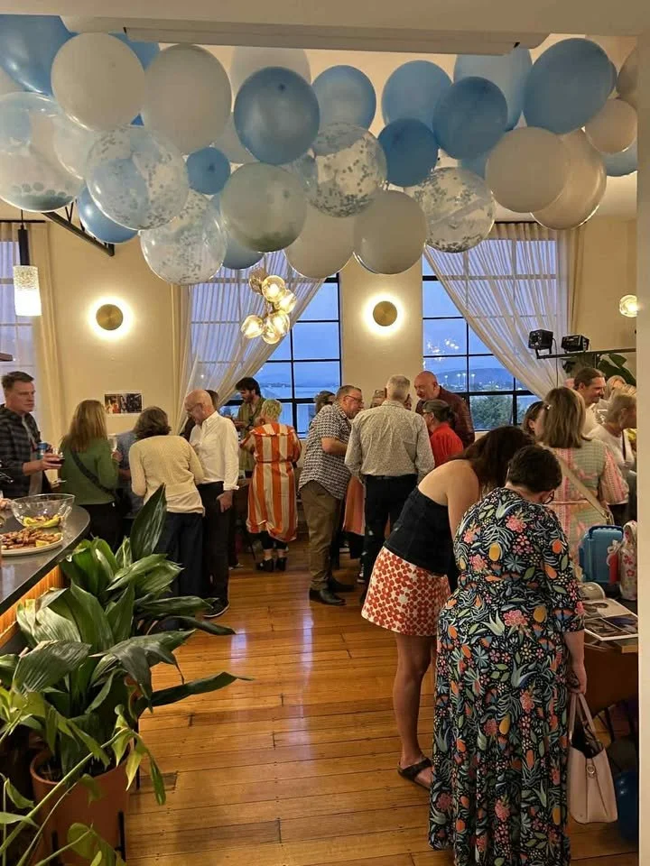 Indoor party with people socializing around tables, large blue and white balloons hanging from the ceiling, and warm lighting. There are windows with curtains, wooden floors, and some plants.