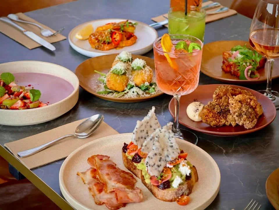 A variety of colorful dishes on a dining table, including a plate of bacon, a crostini with cheese, figs, and chips, fried chicken with a dipping sauce, a pink cocktail with orange peel and mint, green and pink drinks, a fruit salad, and other appetizers and drinks.