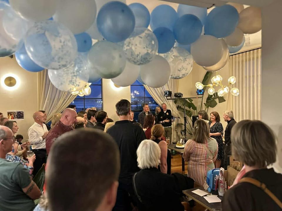 Group of people gathered at an indoor event with large blue and white balloons overhead and a person speaking on a stage.
