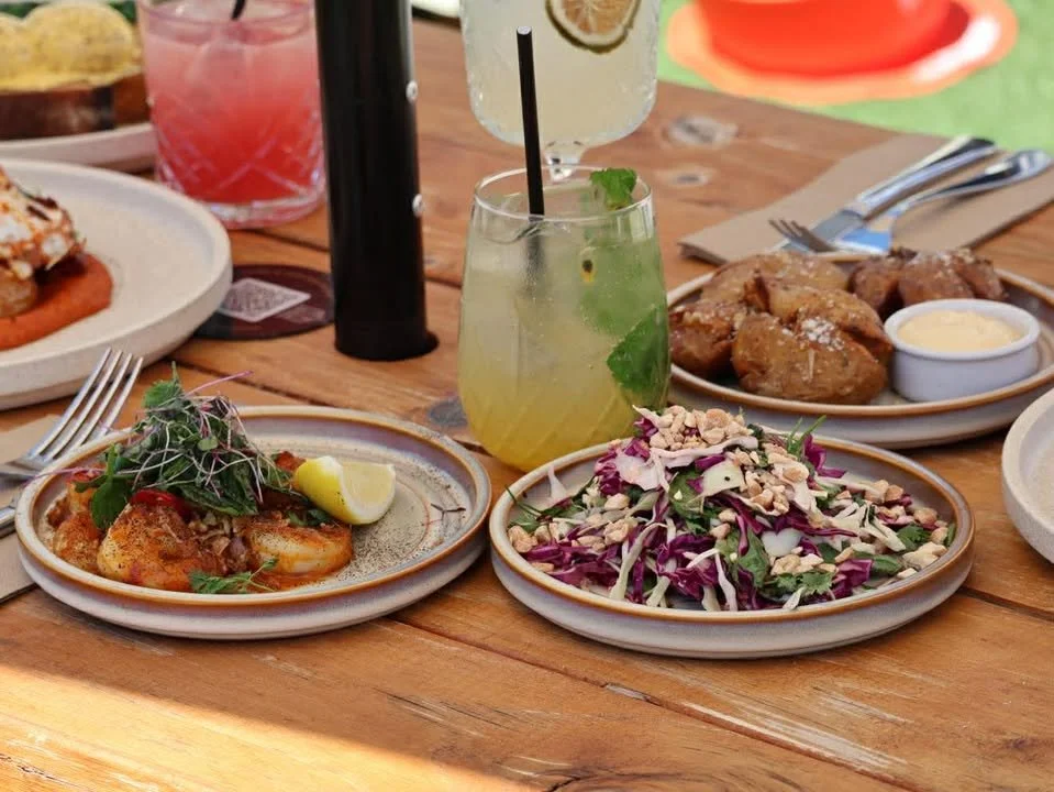 A wooden table with plates of food including shrimp, salad, chicken wings, and drinks.