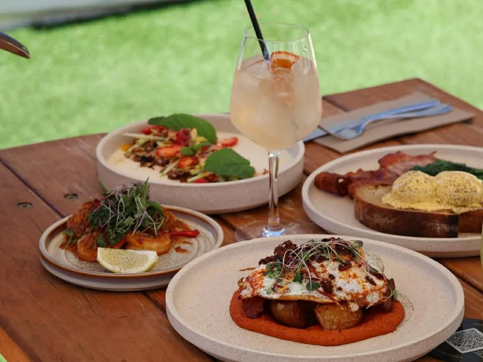 A wooden table with various dishes including a plate of eggs with vegetables, a plate of bacon and toast with eggs, a bowl of salad topped with strawberries and greens, and a small plate of protein with lemon and greens. There is also a glass of a li