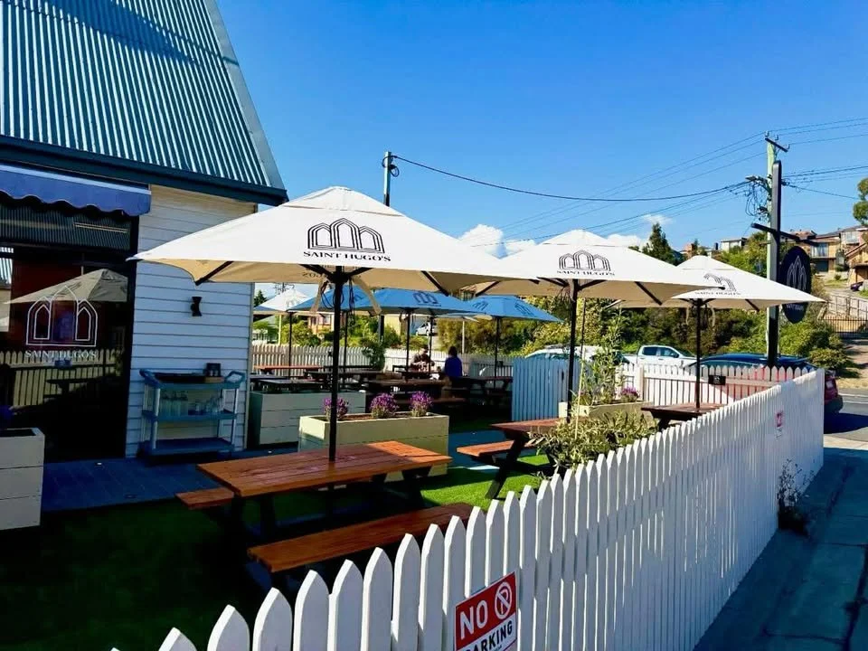 Outdoor seating area of a café with white umbrellas, wooden picnic tables, and a white picket fence under a blue sky.