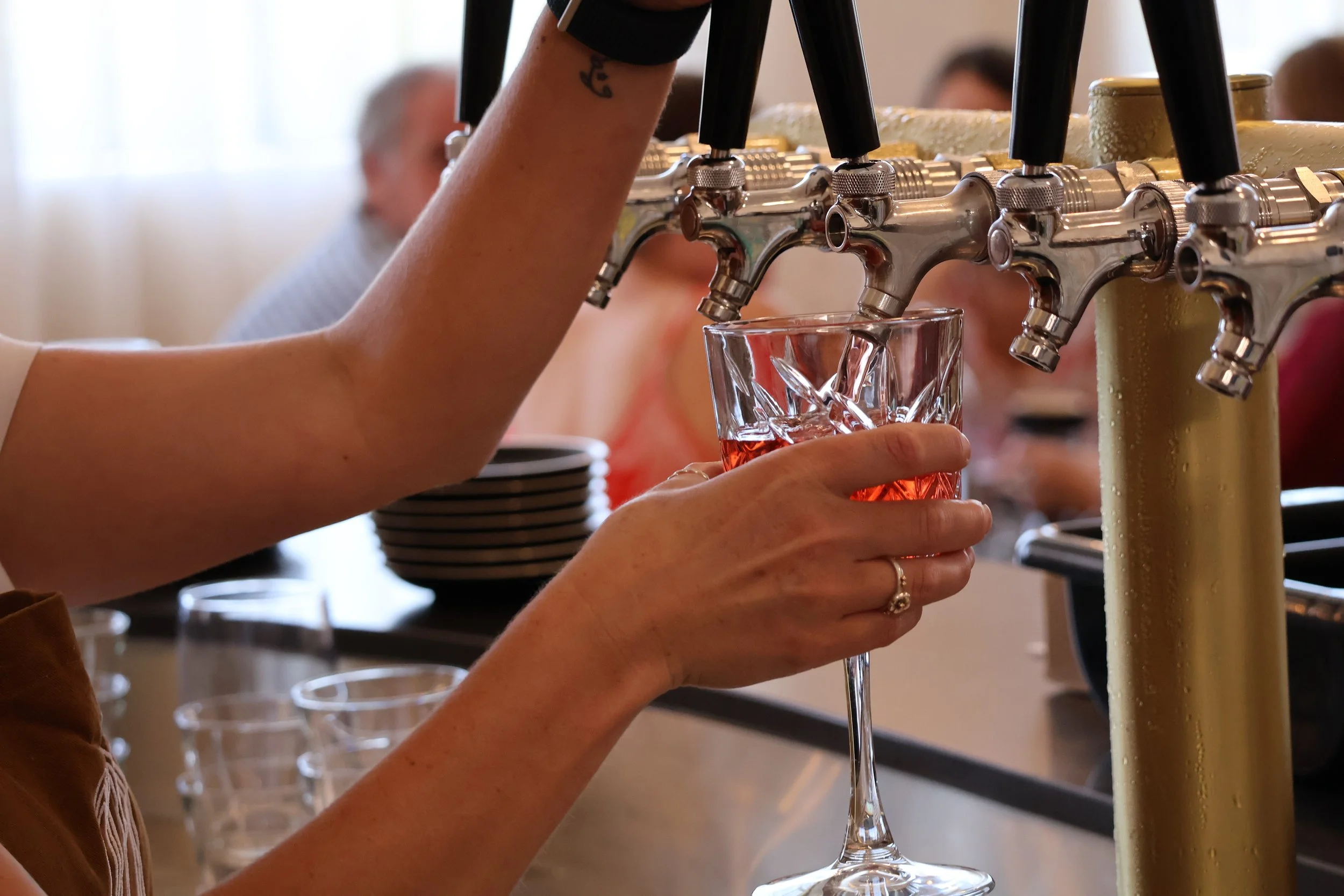 A person filling a glass with beer from multiple taps at a bar or brewery. The person is wearing a ring and has a tattoo on their arm.