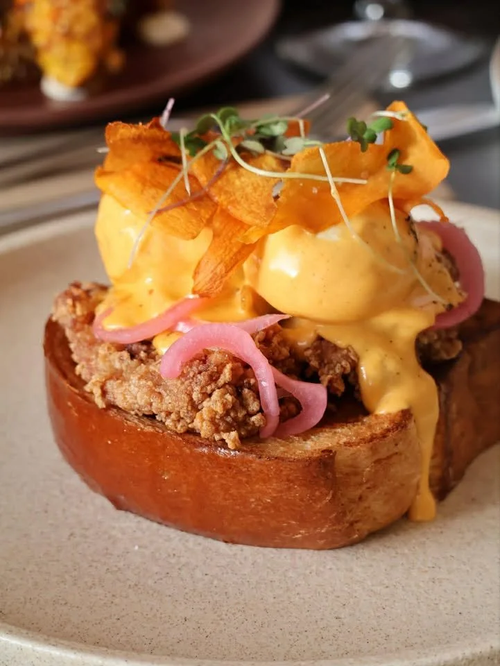A brunch dish featuring a halved toasted bread bowl filled with ground meat, topped with poached eggs, cheese sauce, crispy potato chips, pickled onions, and microgreens.