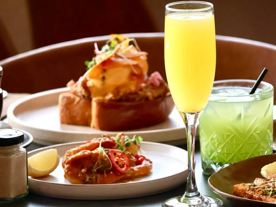 A table with a glass of yellow mimosa, a glass of green drink, a plate with a piece of chicken topped with sliced red chili and herbs, and a small lemon wedge. In the background, a plate with a sandwich or burger filled with vegetables and meat.