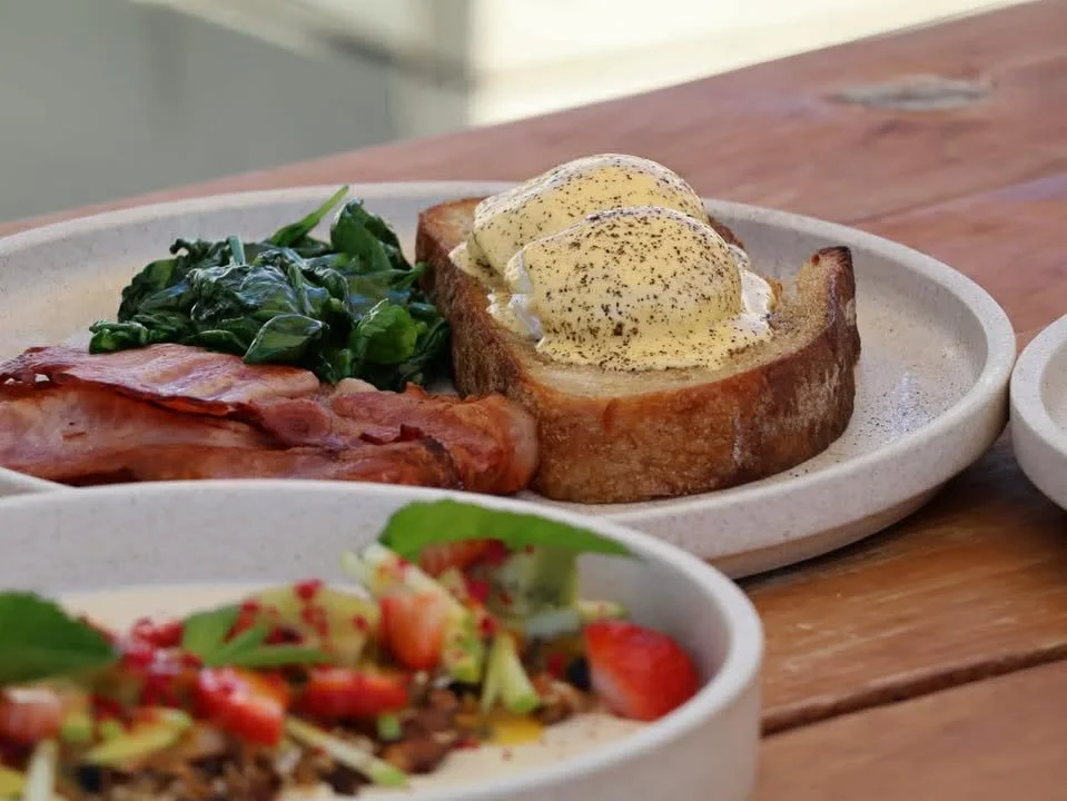 A plate of breakfast featuring eggs Benedict with hollandaise sauce, spinach, crispy bacon, and toasted bread on a wooden table.