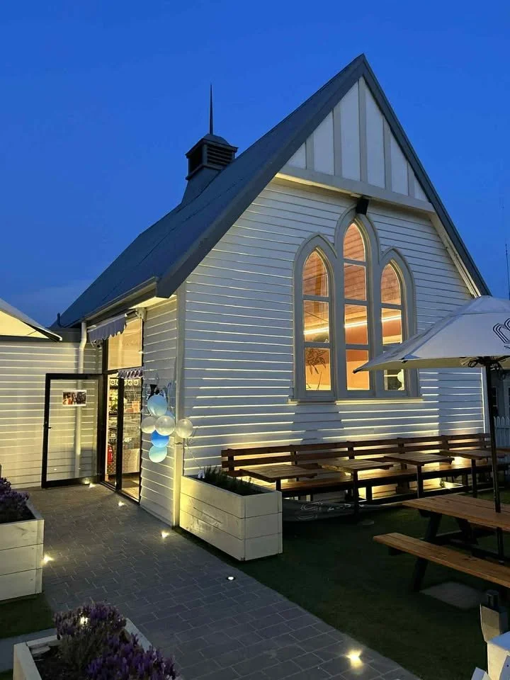 Exterior view of a white church building at dusk with illuminated interior, tall arched windows, a steep gabled roof, and an outdoor seating area with picnic tables and umbrellas.