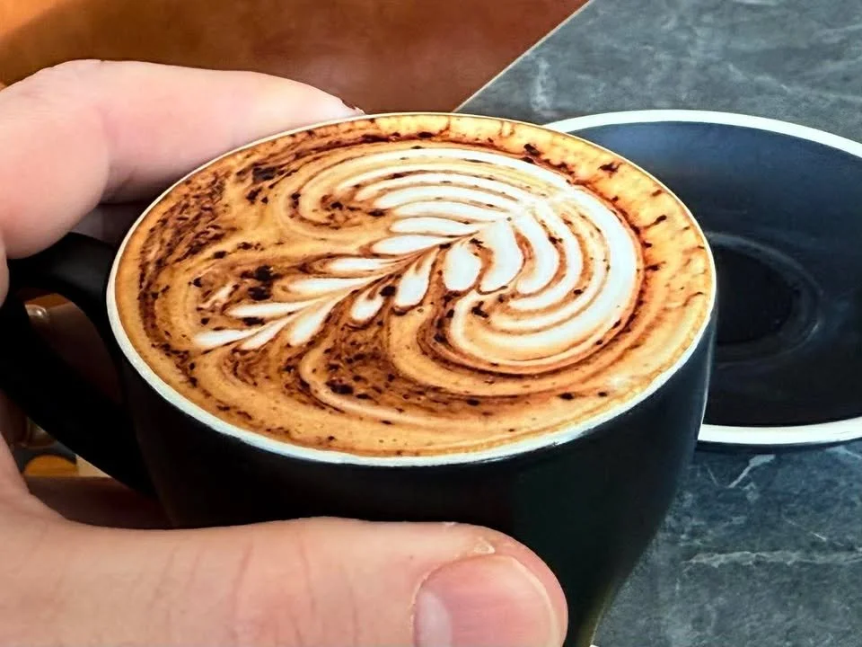 A hand holding a black ceramic mug filled with a cappuccino decorated with chocolate syrup and steamed milk foam art on top, with a matching black saucer nearby.