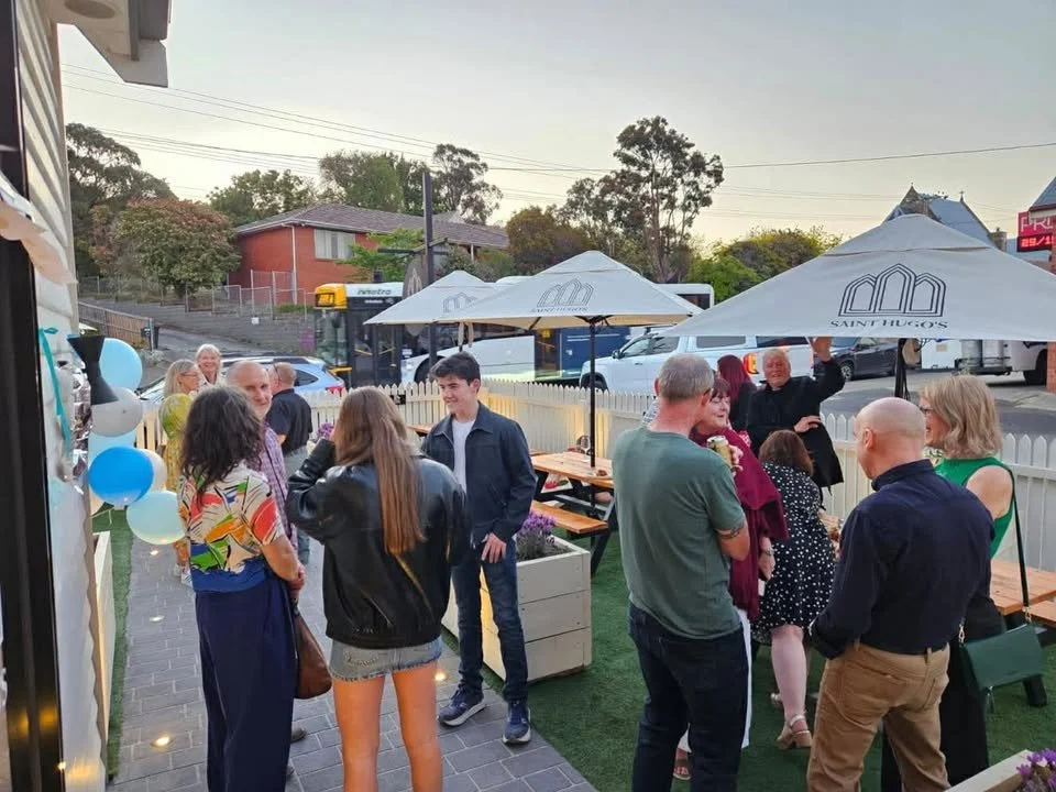 Group of people socializing outdoors at a small gathering under umbrellas, with string lights and balloons decorating the area.