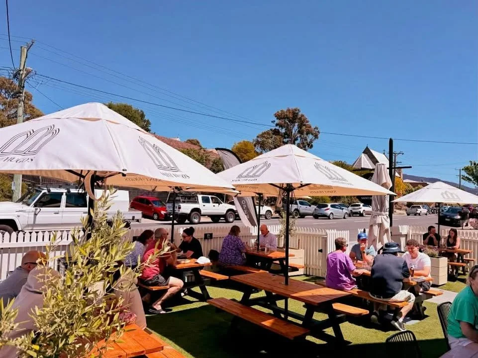 Outdoor patio with people sitting at picnic tables under large umbrellas at a restaurant, with a white picket fence, cars on the street, and trees and buildings in the background on a sunny day.
