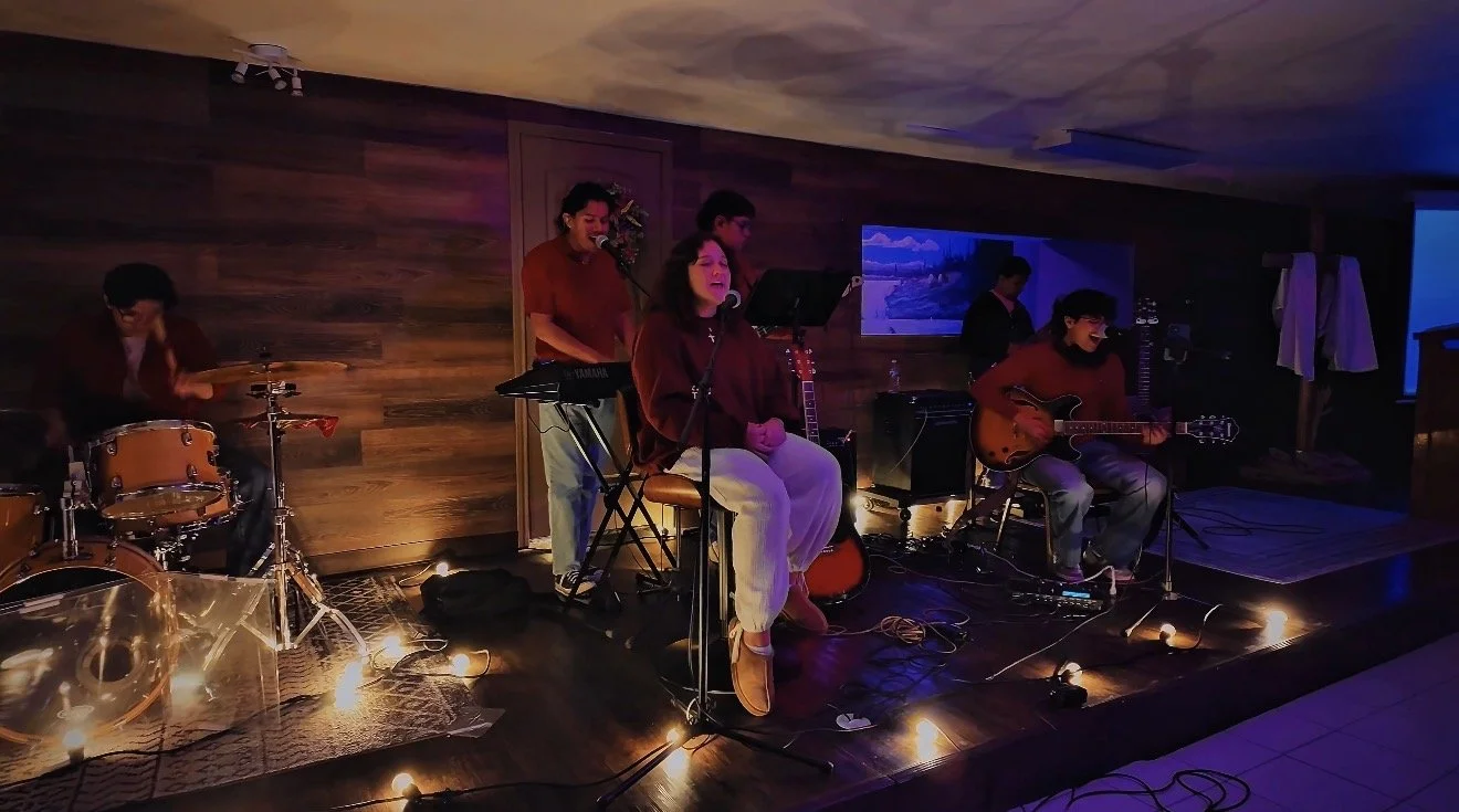 A five-member band performing on a cozy wooden stage with dim lighting, one person singing, two playing guitars, one playing drums, and one possibly playing a keyboard.
