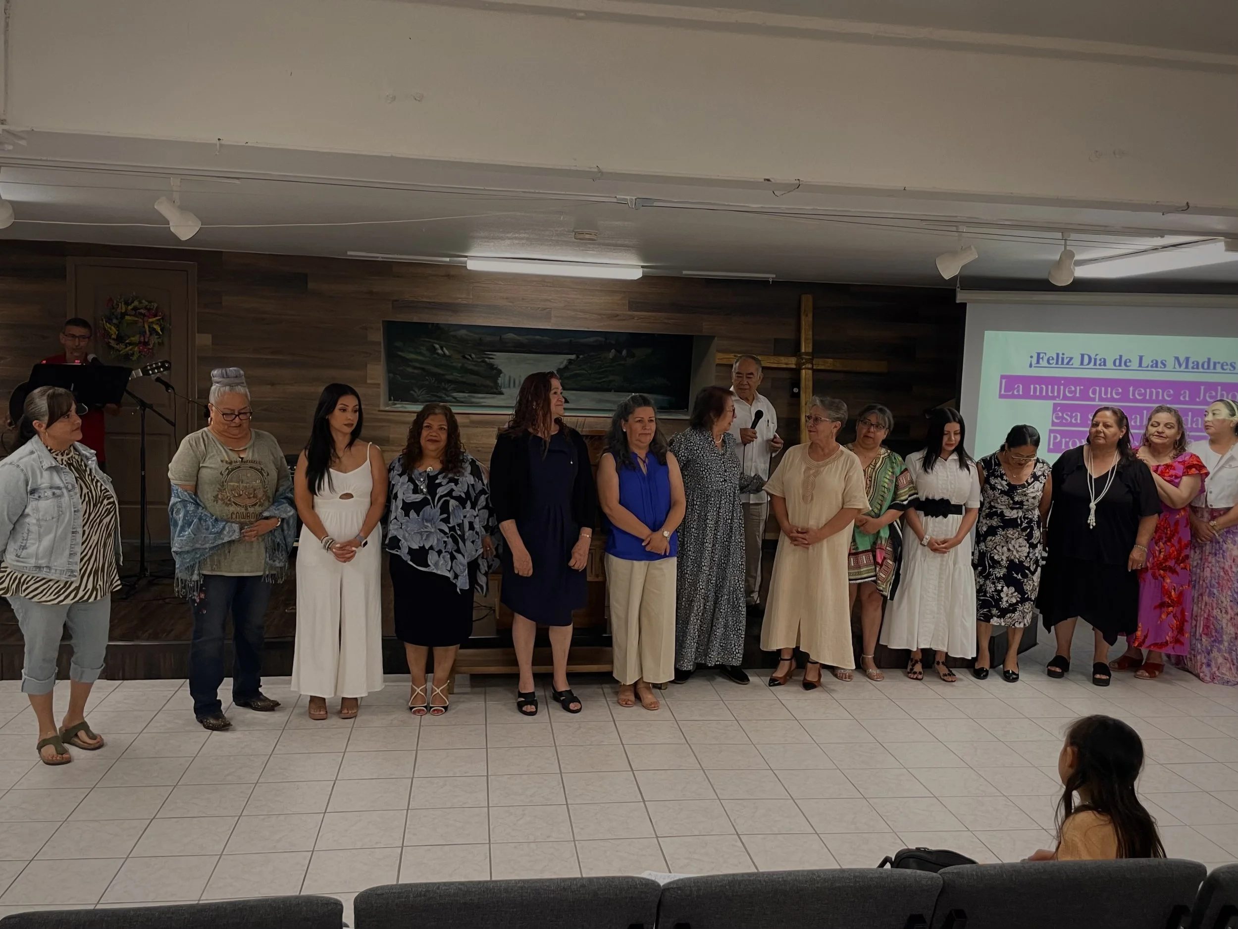 A group of women is standing in a line in a church or community center, with some holding microphones, during a celebration for Mother's Day. There is a large cross on the wall and a screen with a message in Spanish.