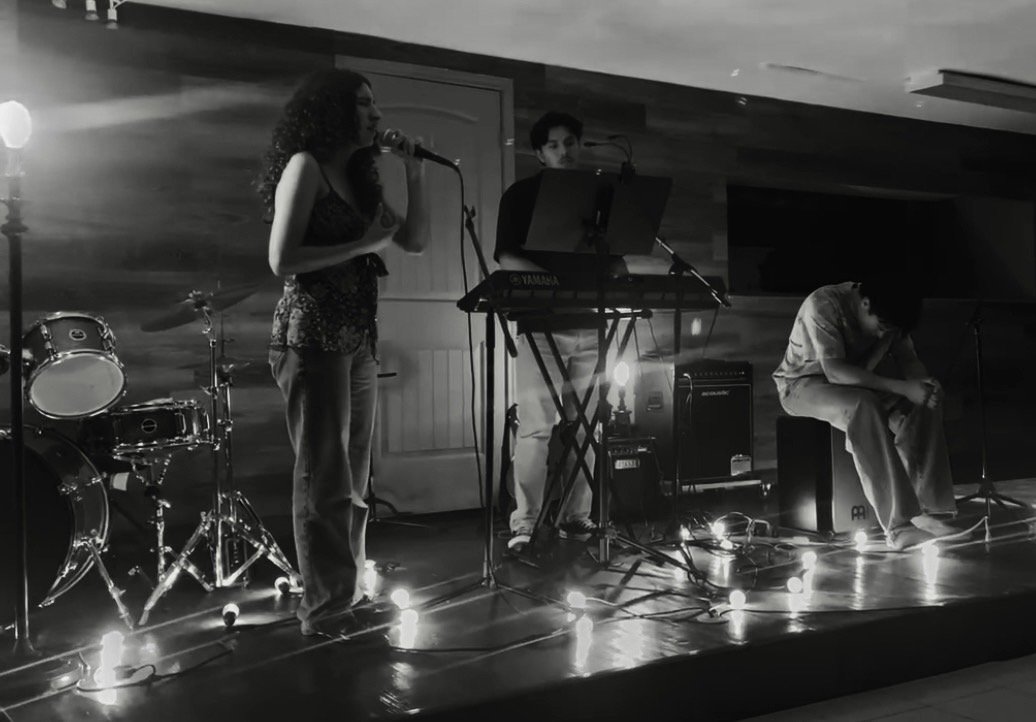 A black and white photo of a musical performance featuring three musicians on stage: a woman singing into a microphone, a man playing keyboard, and another man sitting on a box, possibly resting or preparing. There is a drum set on the left, and string lights on the floor enhancing the stage ambiance.