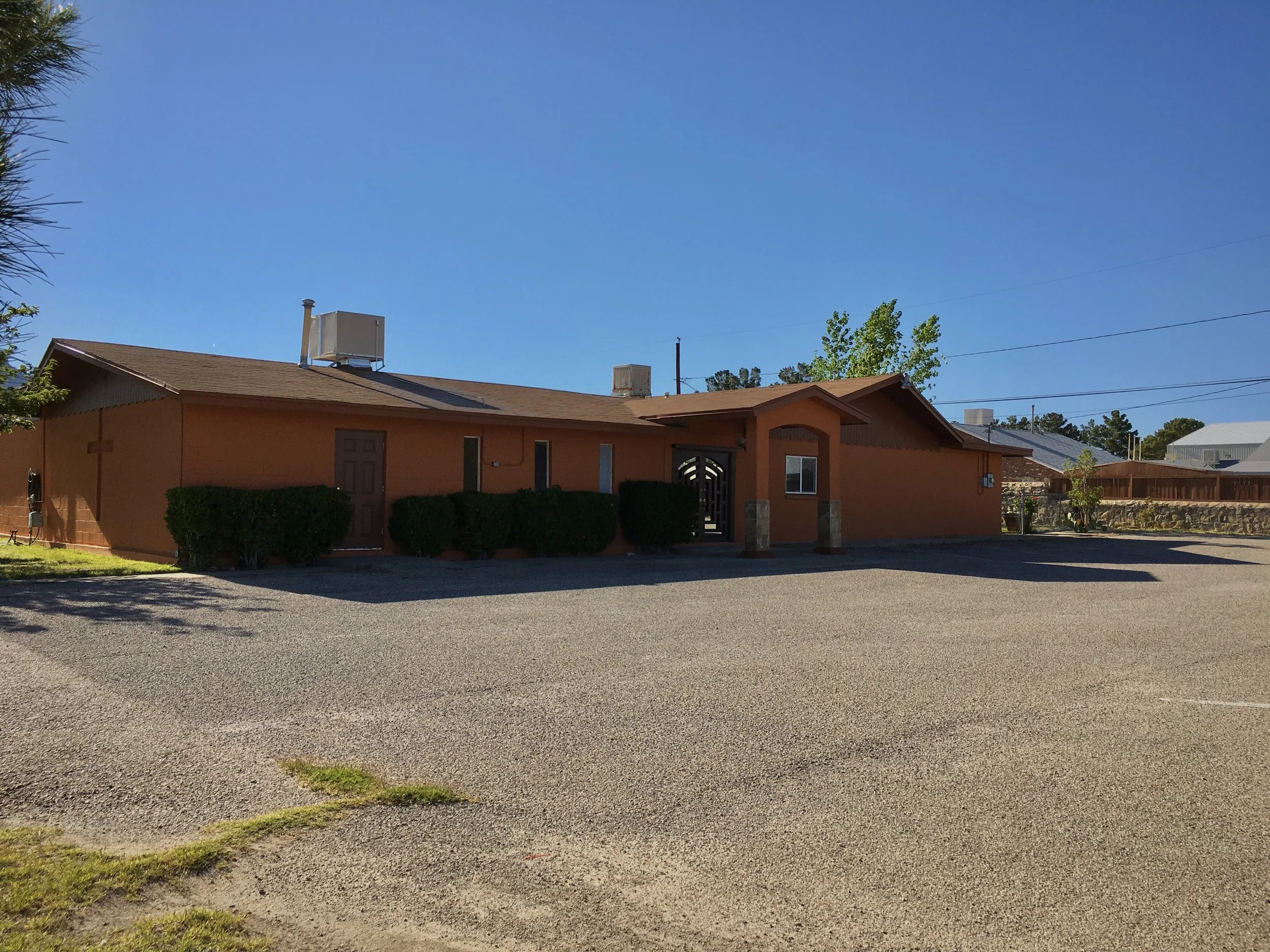 A single-story orange building with a brown roof and a gated entrance, surrounded by a gravel parking lot and some small bushes under a clear blue sky.