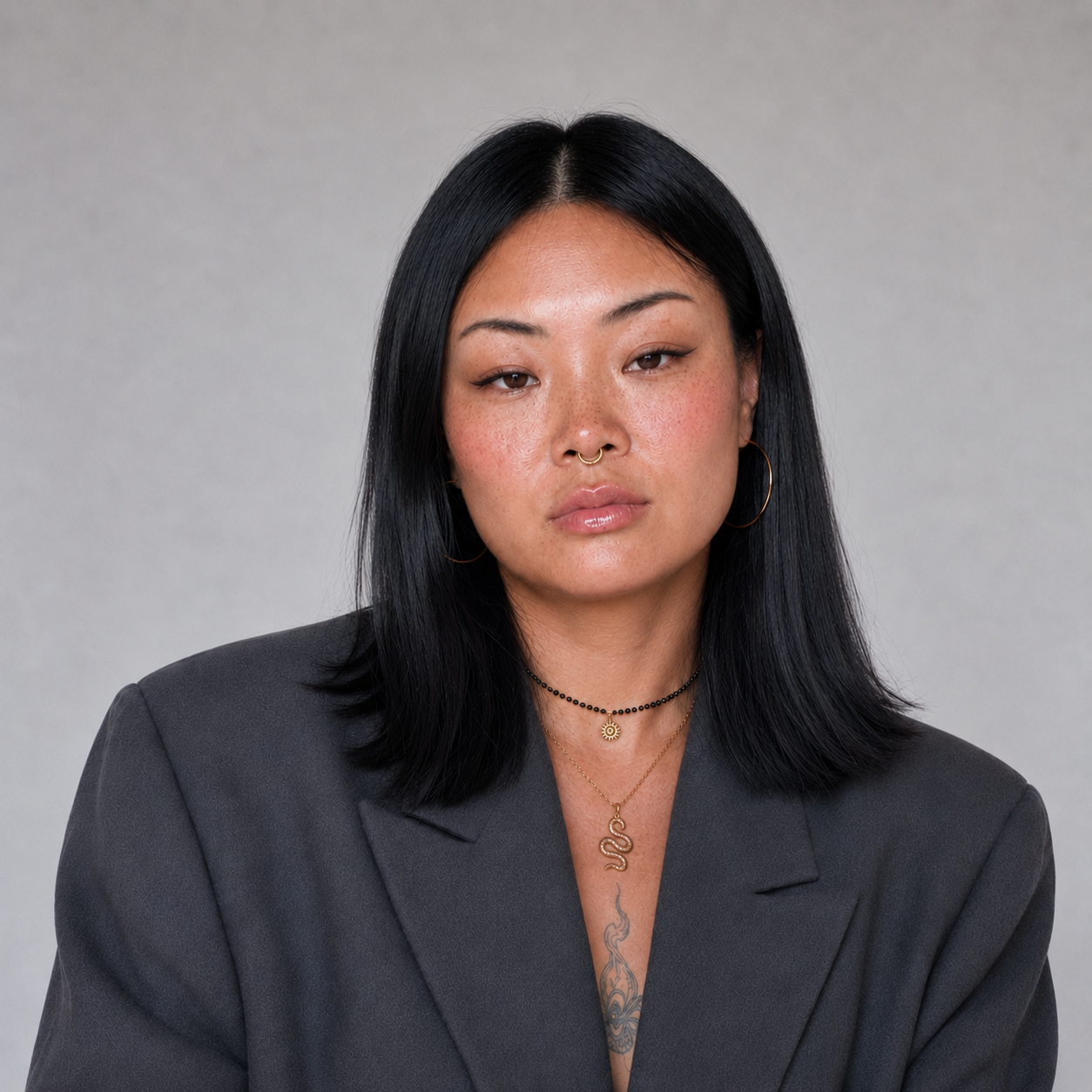 Portrait of an Asian woman with long black hair wearing a dark blazer, multiple necklaces, earrings, and a septum piercing, against a plain gray background.