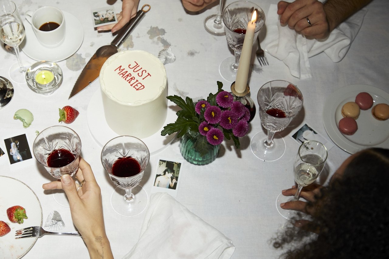 Intimate wedding table scene with a “just married” cake, red wine, polaroids, candles and hands — modern event styling aesthetic