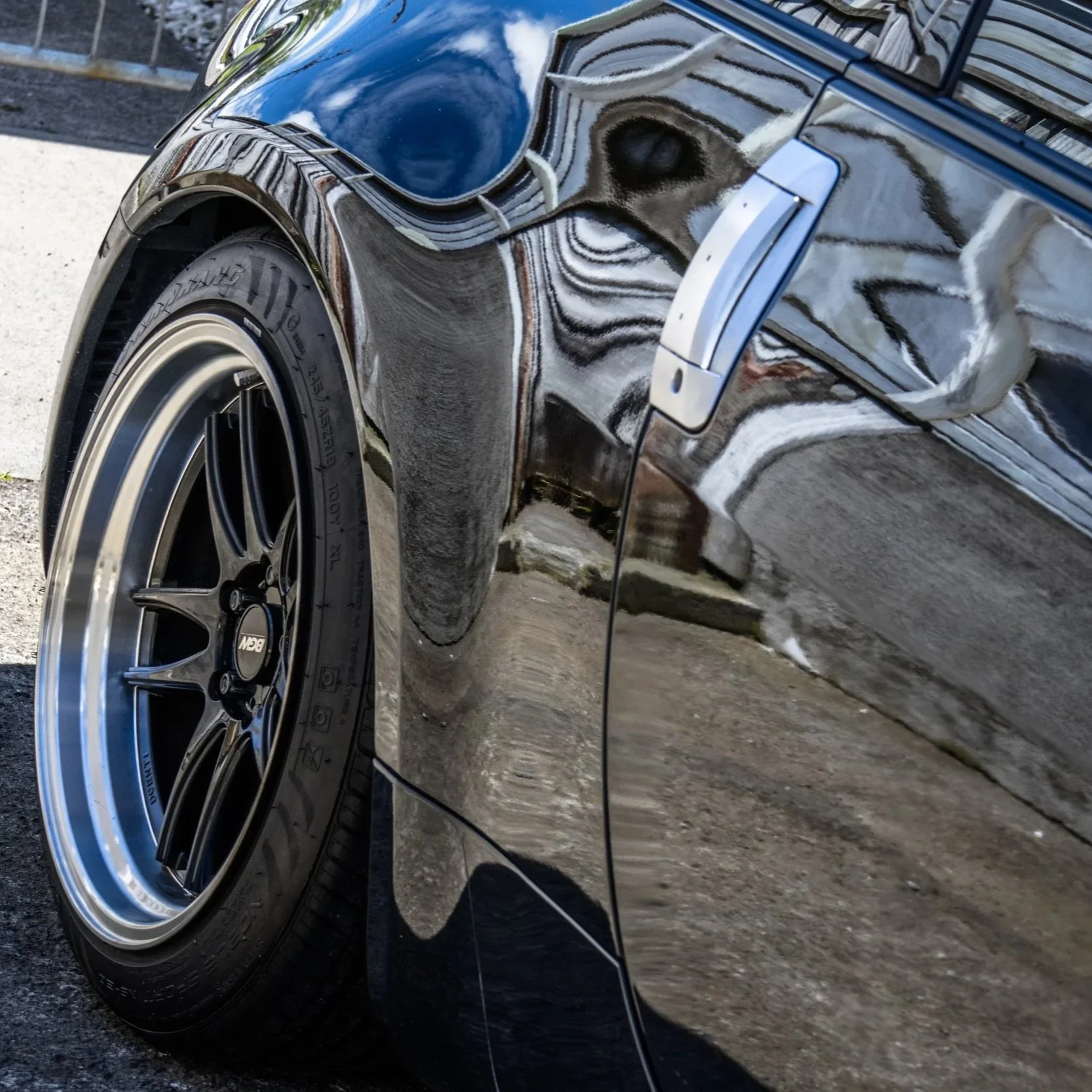 Close-up of a black car's front left wheel and side mirror, showing reflections on the car's shiny surface.