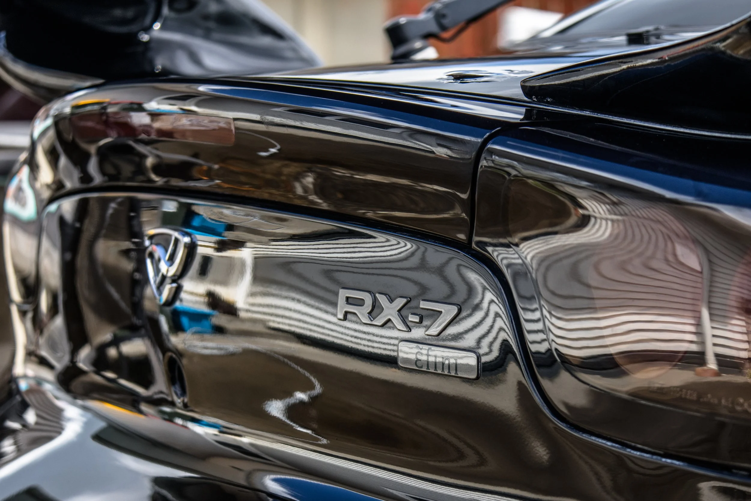 Close-up of a black Toyota RX-7 sports car, showing the rear badge and part of the tail light.