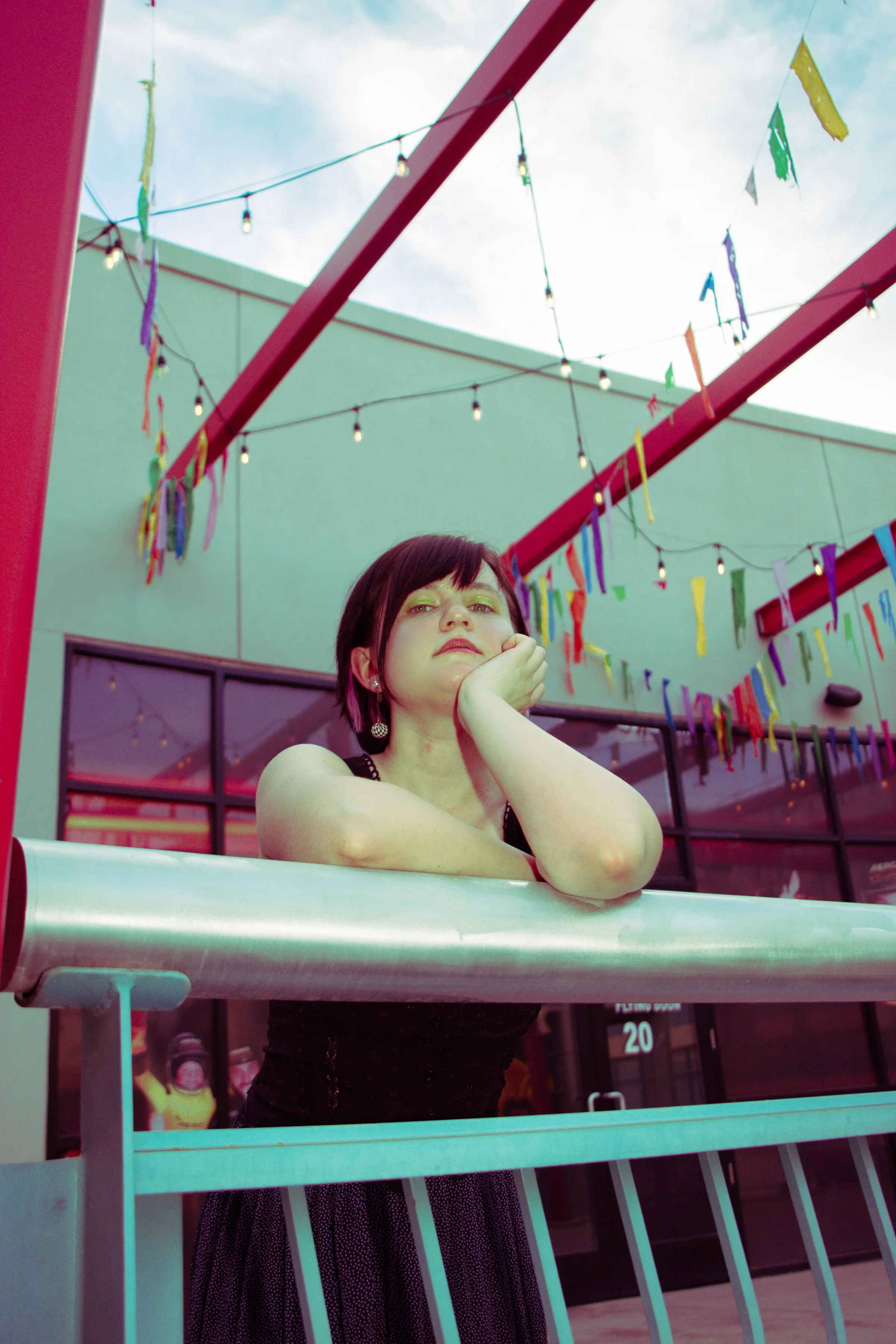 A woman with short dark hair and earrings leaning on her hand, standing behind a white bar railing at an outdoor event decorated with colorful streamers and string lights.