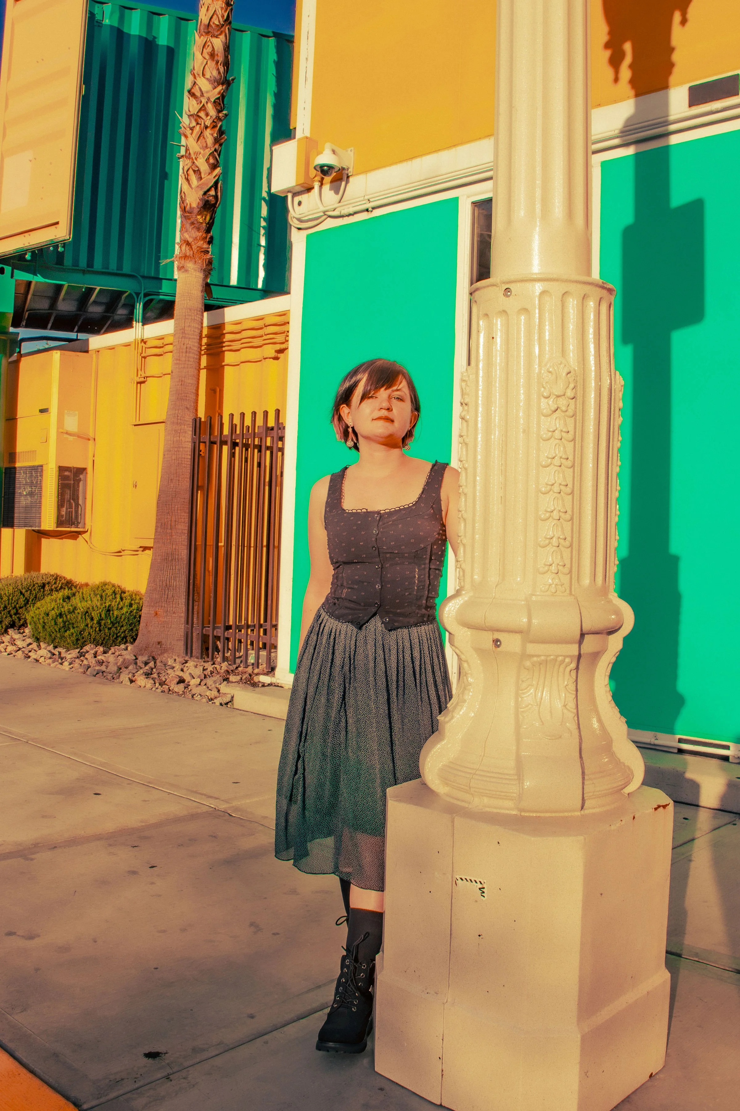 A woman with short hair and earrings leaning against a decorative cream-colored lamppost on a sidewalk, with colorful buildings and a tree in the background, under bright sunlight.