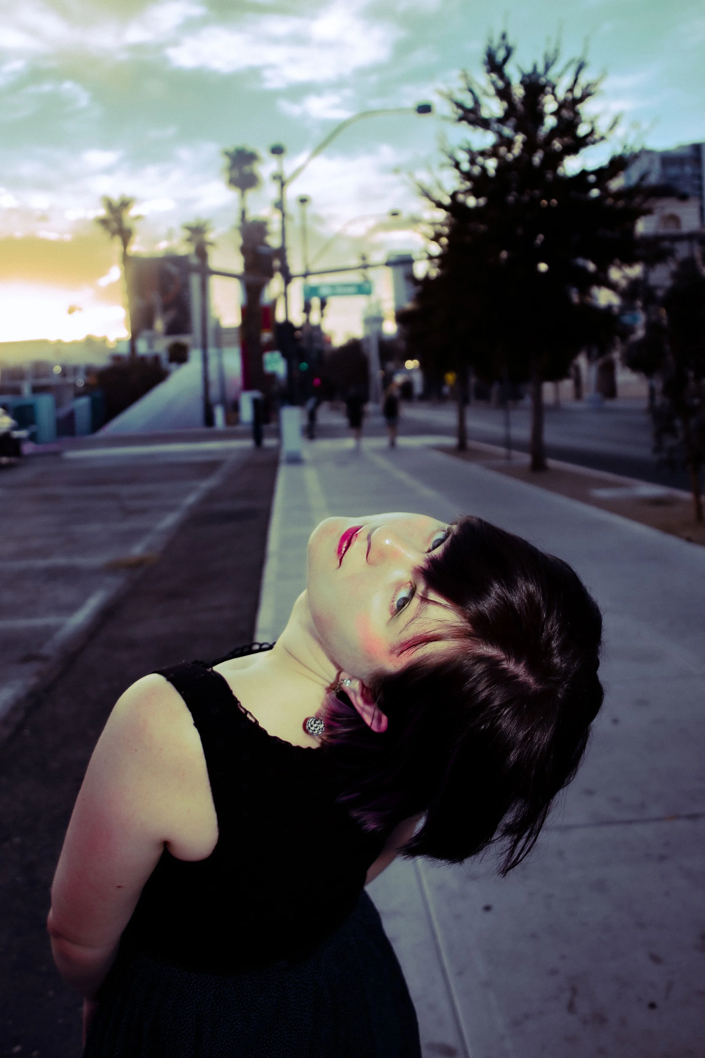 A woman with dark hair and light skin leaning back with her head tilted upward on a city sidewalk, with trees and streetlights in the background during sunset.