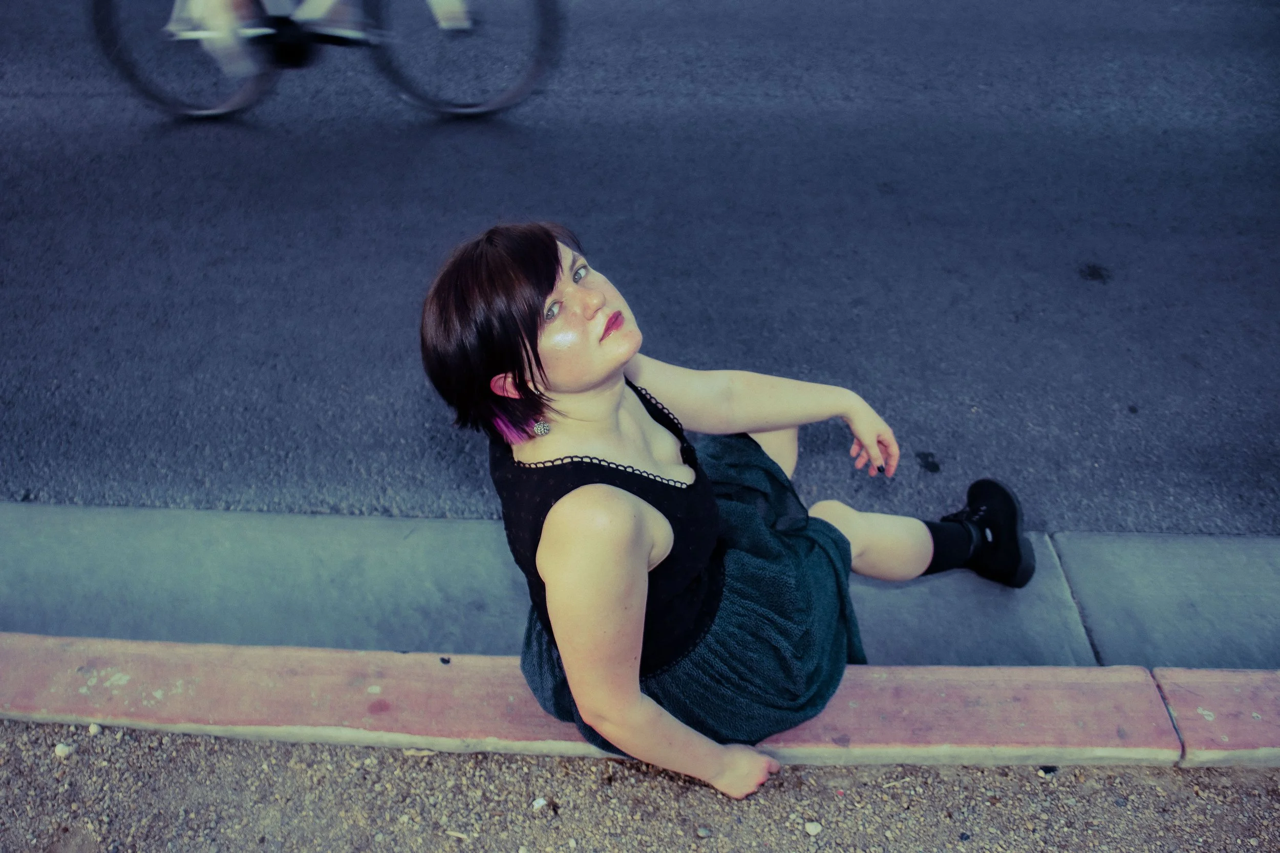 A woman with short dark hair and pink highlights, wearing a black dress and black boots, sitting on a curb at night, gazing upward.