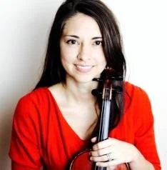 Woman with long dark hair wearing a red top, holding a violin, smiling at the camera.