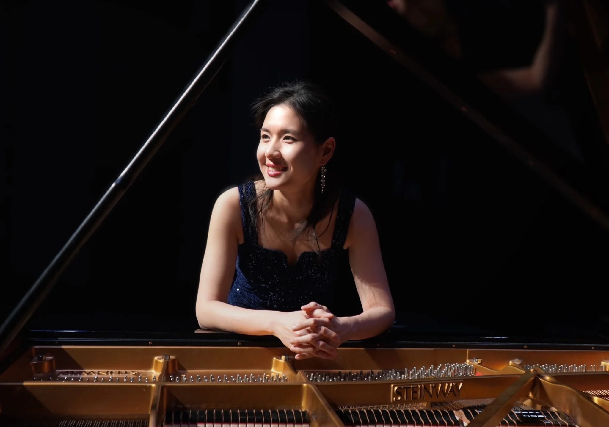 A woman in a navy blue dress with earrings, smiling, leaning on a grand piano with her arms crossed on top of it.