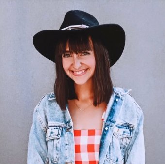 Young woman smiling wearing a black wide-brim hat, denim jacket, and red and white checkered top, standing against a plain background.