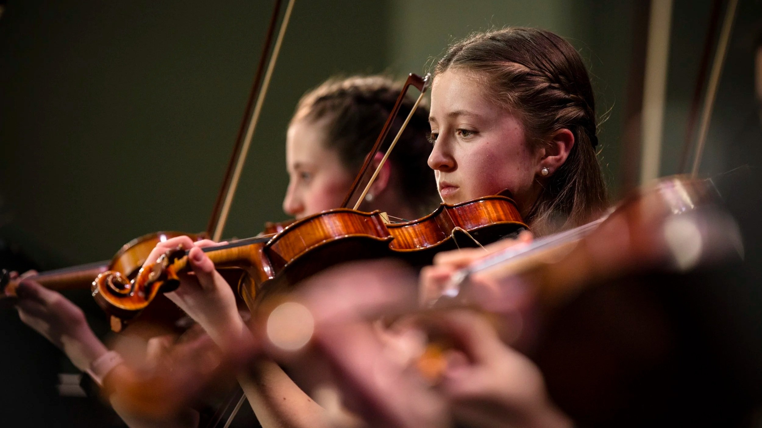 Young women playing violins in an orchestra, focused on their instruments during a performance.