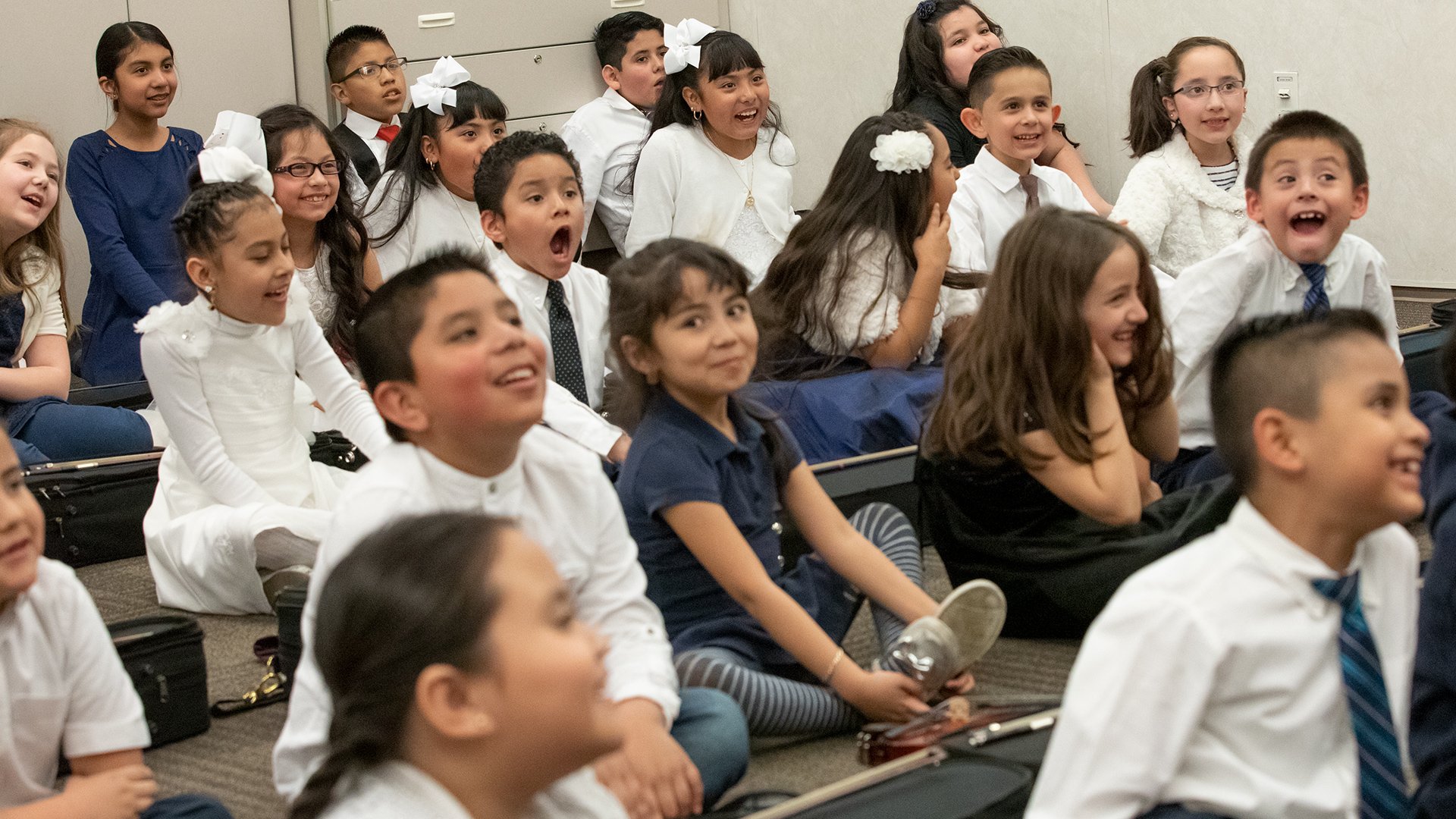 Group of young children sitting on the floor and listening attentively, some smiling and laughing, in a classroom or educational setting.