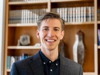 Young man in a dark blazer and dress shirt smiling in a library or office setting with shelves of books and decorative items in the background.