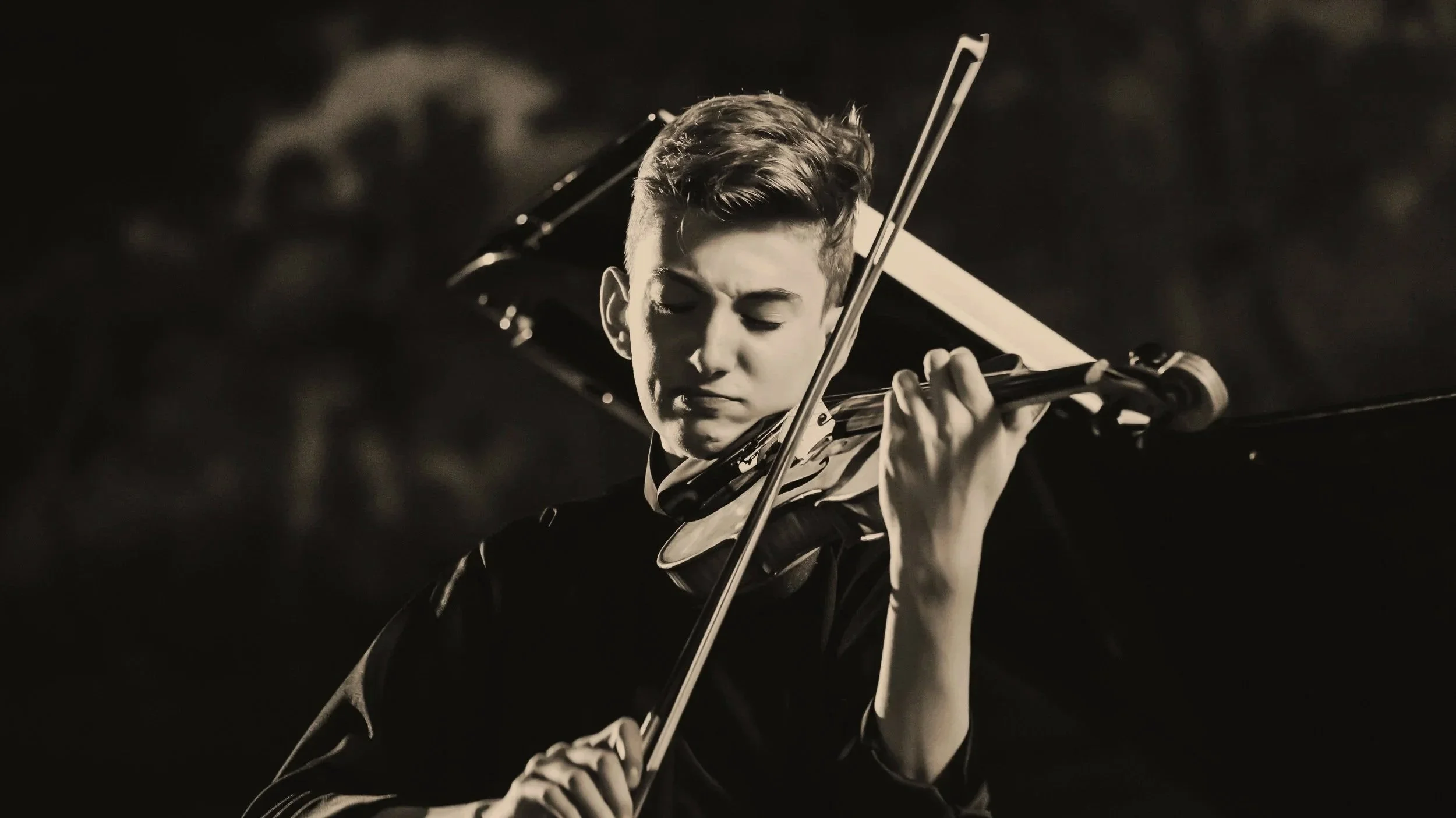 A young man with short, styled hair playing the violin with his eyes closed, focused on his music in a black and white photograph.