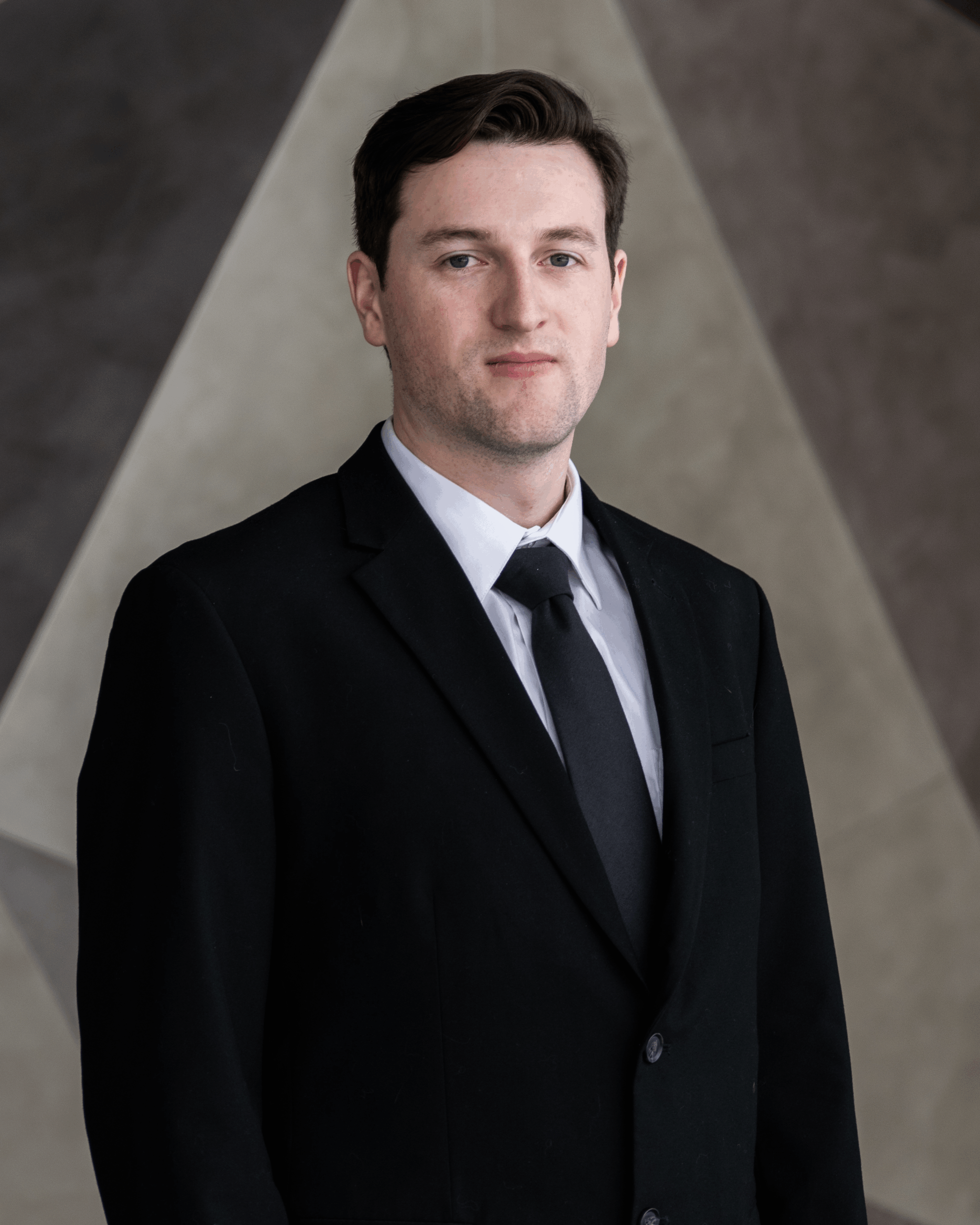A man in a black suit, white shirt, and black tie standing in front of a geometric background.
