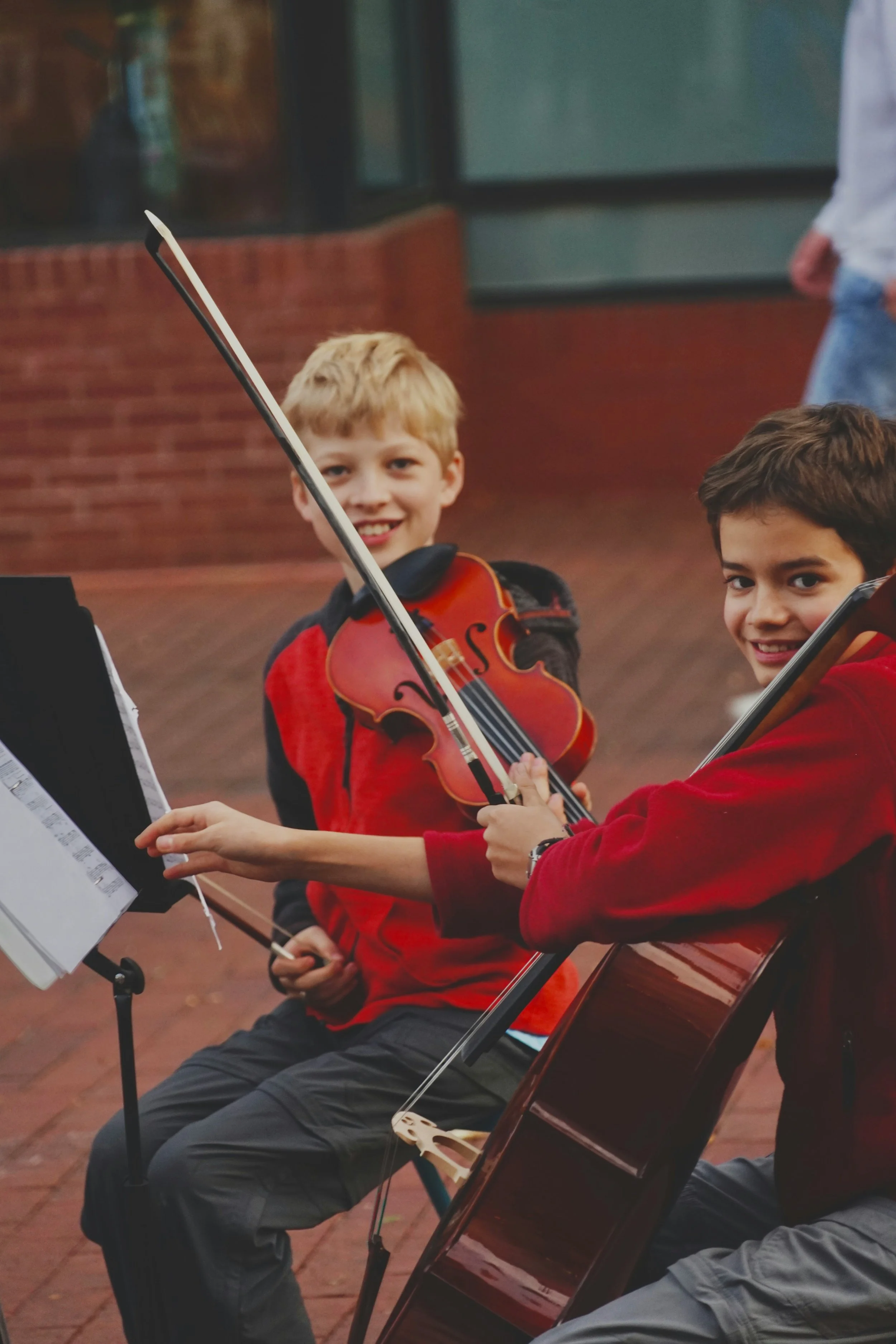 Two young boys playing violins in an outdoor setting, seated in a music recital, with sheet music on stands, and a brick wall in the background.