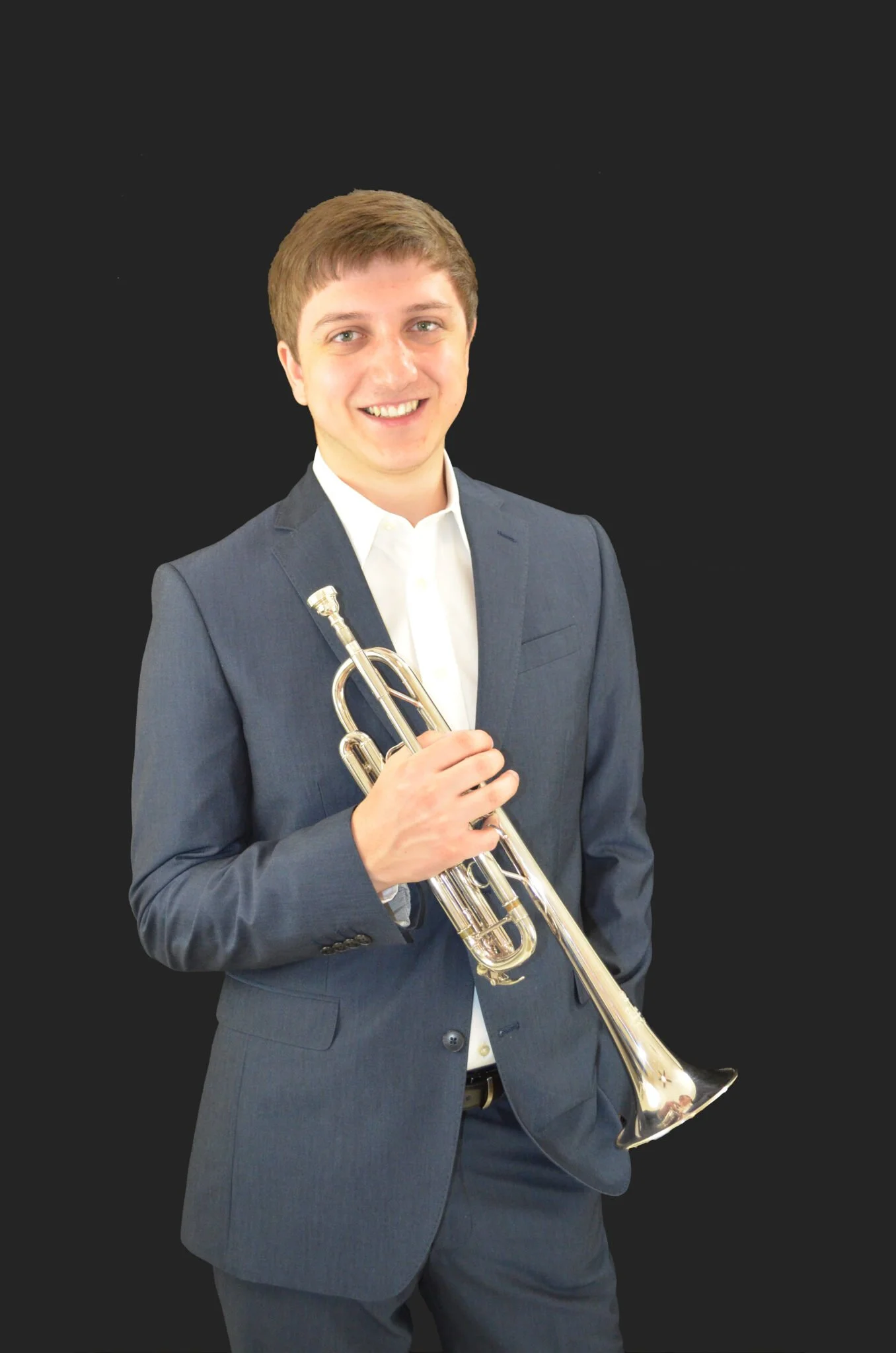 A young man in a dark gray suit and white shirt holding a trumpet, smiling, against a dark background.