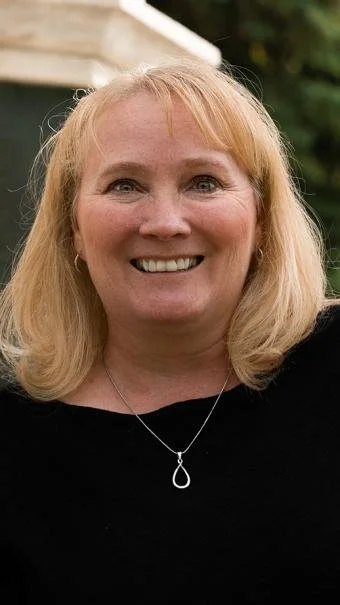 Close-up of a smiling woman with blonde hair, wearing a black top and silver necklace, standing outdoors with a blurred background.