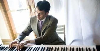 Man in a suit playing a piano in a well-lit room with large windows and white curtains.