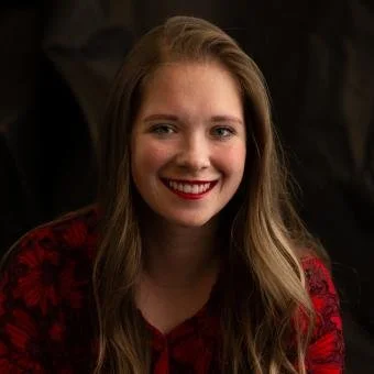 A young woman with long, wavy hair and a bright smile, wearing a red patterned top, against a dark background.