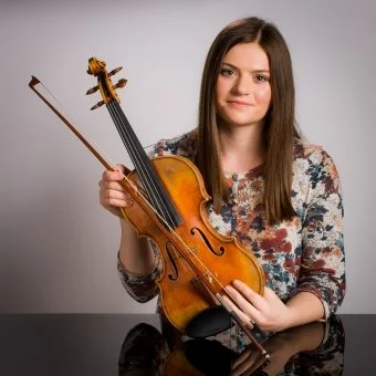 Young woman holding a violin and bow, smiling at the camera, against a plain background.