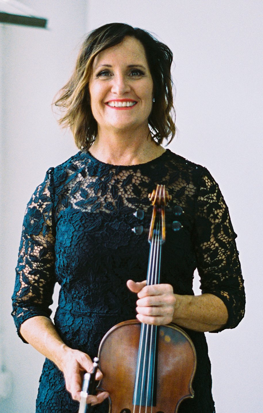 A woman with shoulder-length wavy brown hair and light skin smiling, holding a violin in her left hand, dressed in a black lace dress, standing indoors with a plain background.