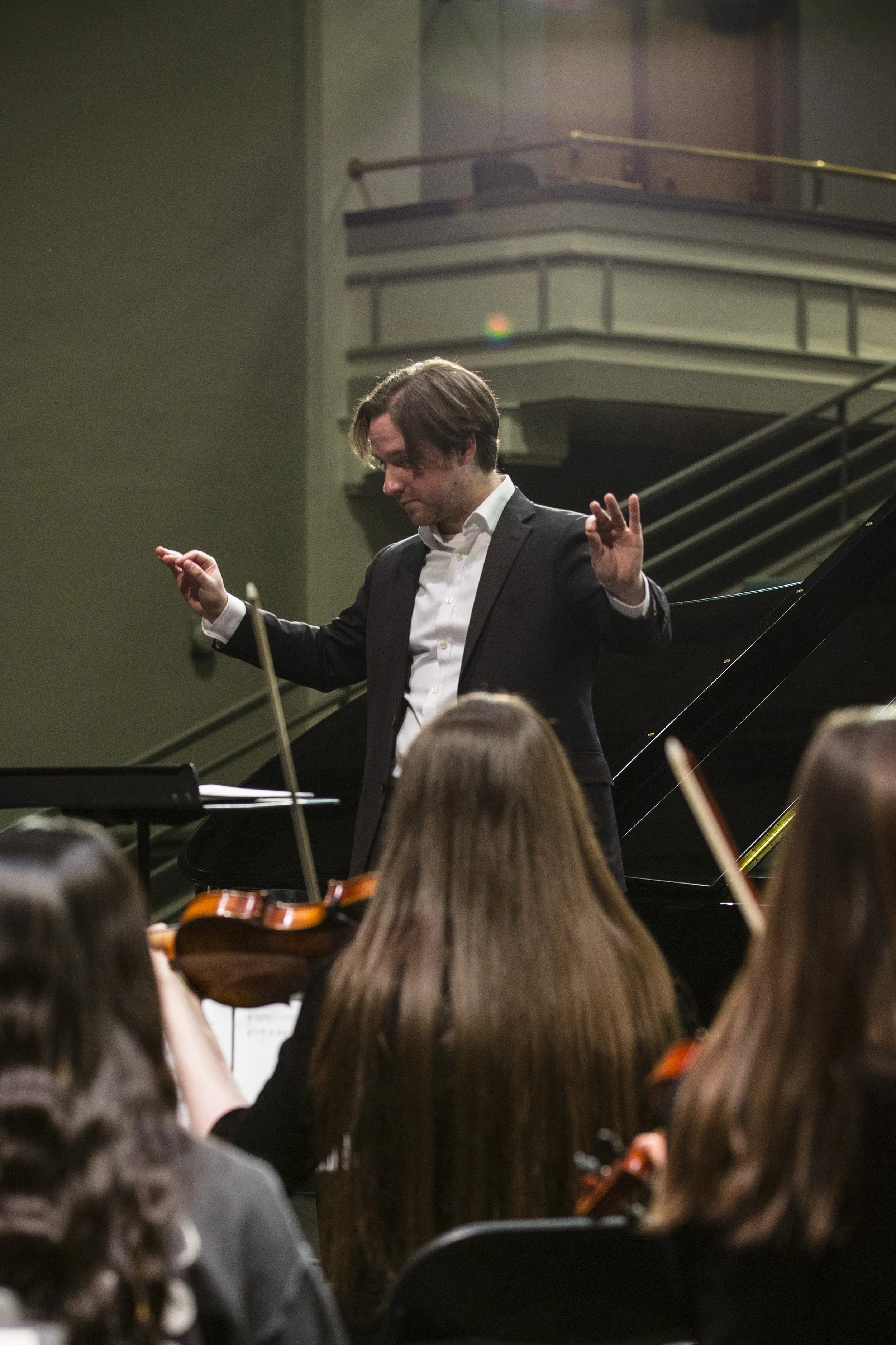 A conductor in a dark suit and white shirt leading an orchestra, with the audience and musicians in the foreground, in a concert hall with balconies.