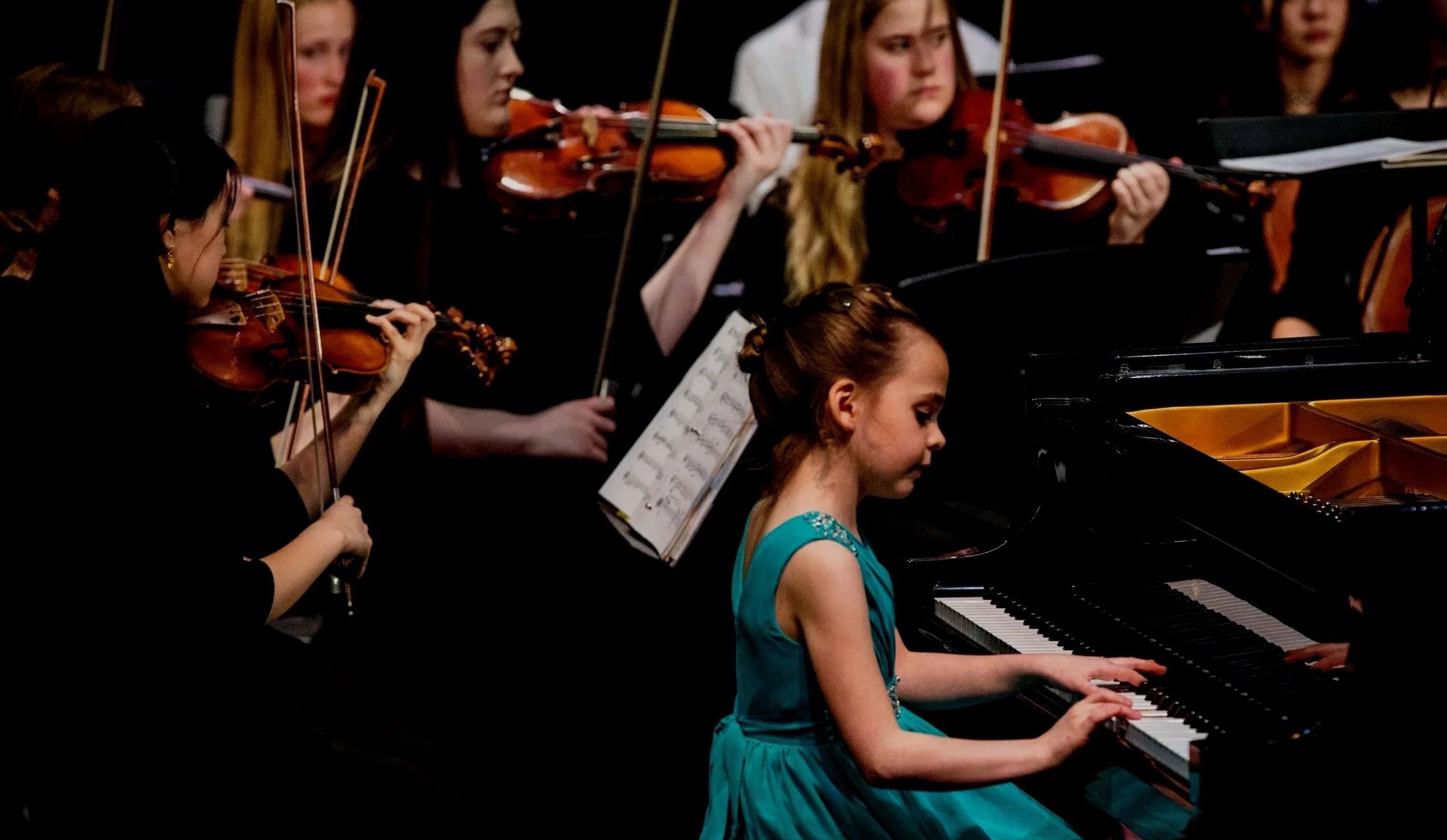 Young girl in a blue dress playing the piano during an orchestra performance, with several women playing violins behind her.