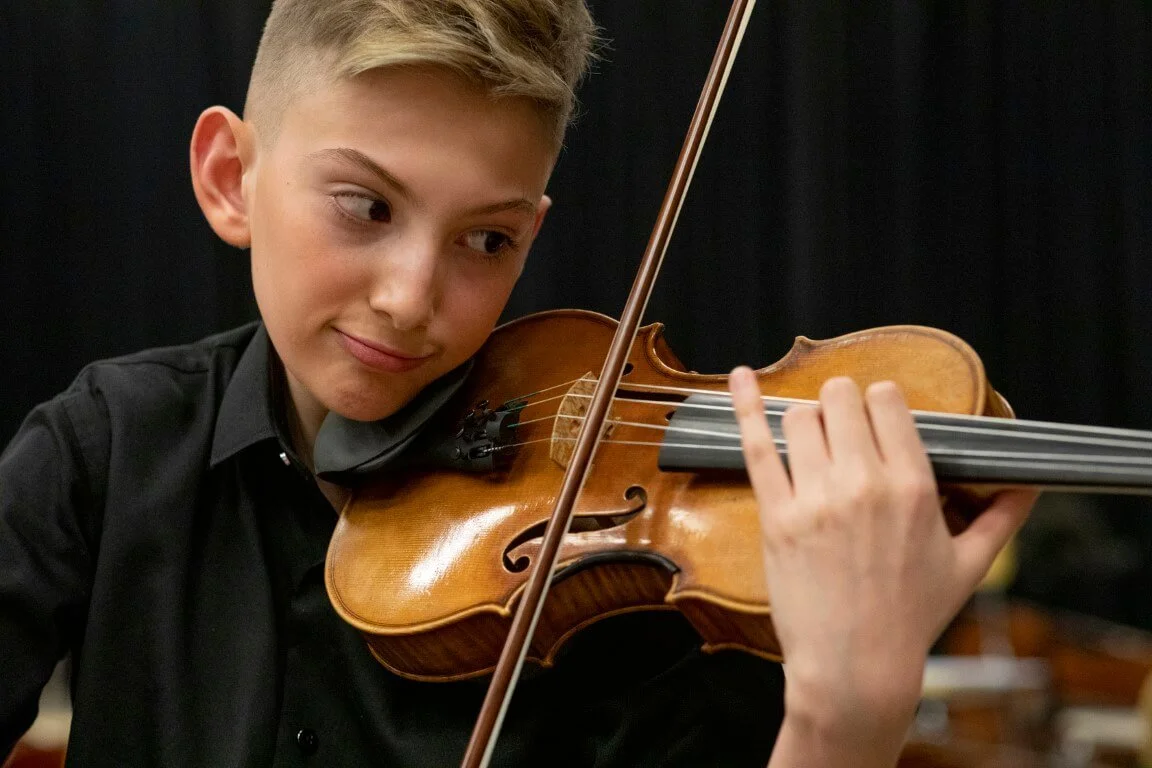 Young boy with short blonde hair playing a violin, wearing a black shirt, against a dark background.