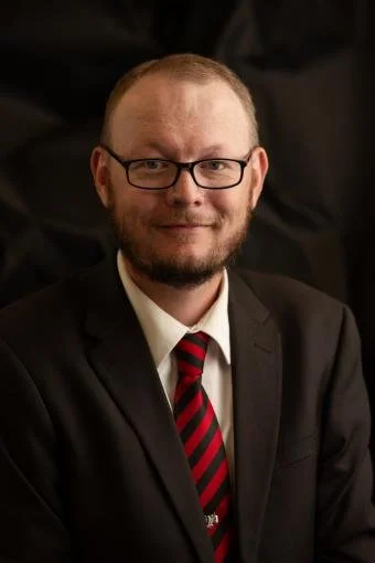 A man with glasses and a beard wearing a suit and red striped tie, smiling at the camera.