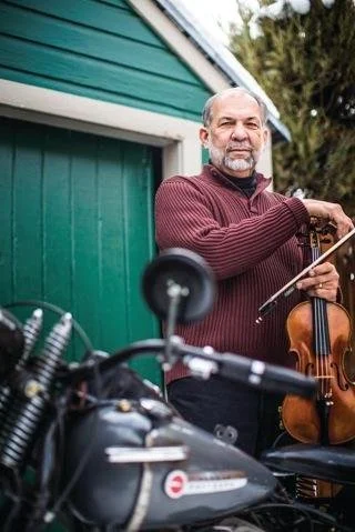Older man with gray hair and beard, wearing a maroon sweater, holding a violin and bow, standing outside next to a green building with a motorcycle in the foreground.